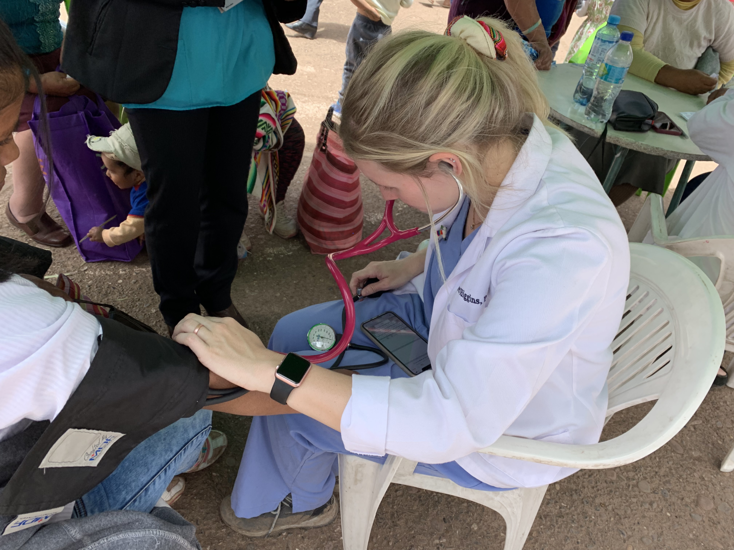 Female healthcare worker in white coat measuring blood pressure of a seated person using a sphygmomanometer and stethoscope outdoors.