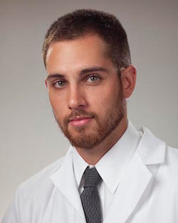 Portrait of a young man with brown hair and beard wearing a white coat and a gray tie against a neutral background.