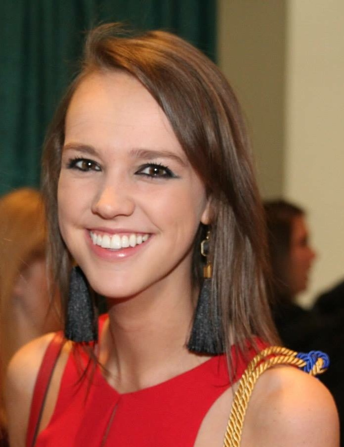 Smiling young woman with straight brown hair wearing black tassel earrings and a red sleeveless top.