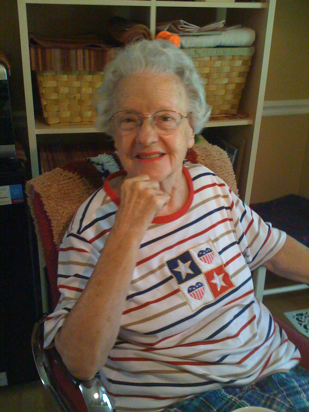 Elderly woman with glasses sitting in a chair, smiling, wearing a striped shirt with patriotic star and flag patches.