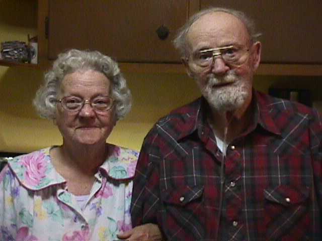 Elderly couple smiling and sitting together indoors, woman with curly gray hair and floral blouse, man with glasses, gray beard, and plaid shirt.
