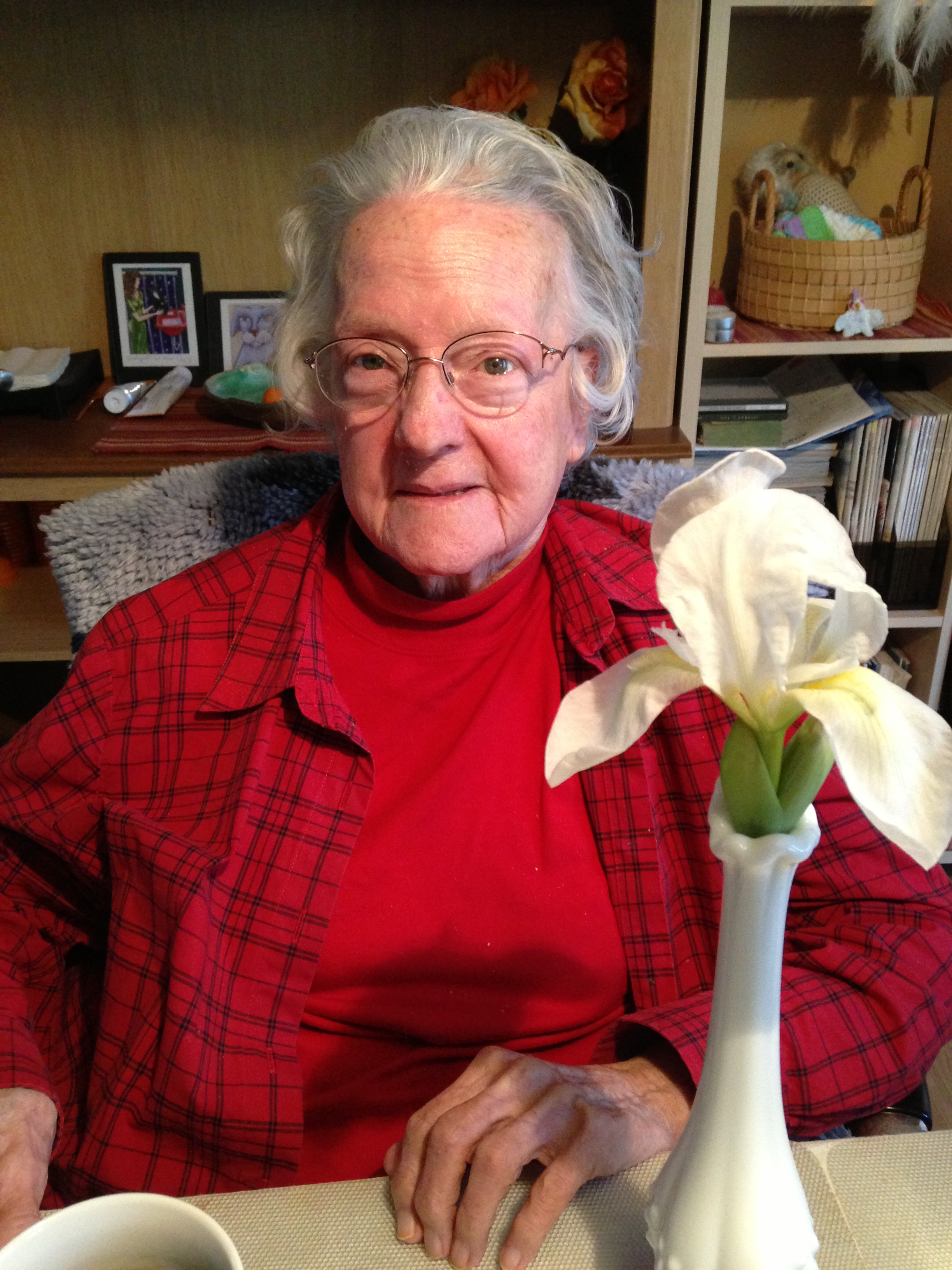 Elderly woman with glasses wearing a red plaid shirt and red turtleneck sitting at a table with a white flower in a vase in front of her.