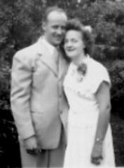 Black and white photo of a smiling couple standing close together outdoors with trees in the background.