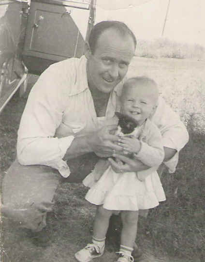 Black-and-white photo of a man crouching and smiling next to a young child holding a small kitten outdoors near a vehicle.