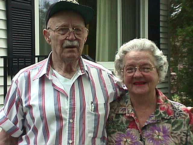 Elderly man wearing a striped shirt and cap standing next to an elderly woman in a floral blouse, both smiling.