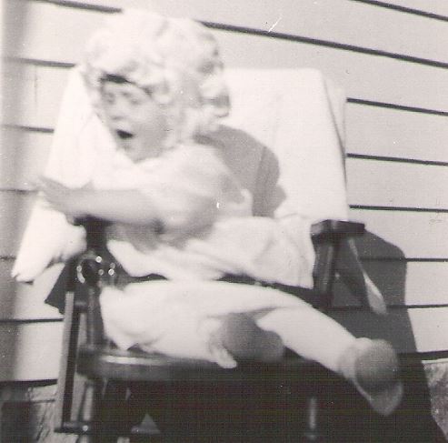 Black and white photo of a young child in a bonnet and dress sitting on an outdoor chair, with one leg extended and an expression of surprise or excitement.