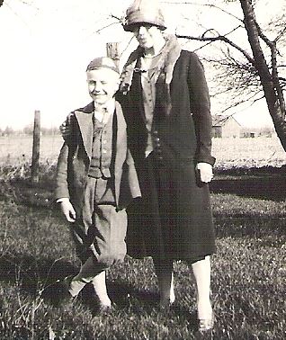 Black and white photo of a woman and a boy standing on grass outdoors, both dressed in vintage clothing and smiling.