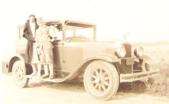 Vintage photo of two women standing beside a classic 1920s car on a dirt road.