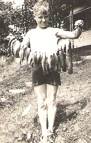 Young boy standing outdoors holding a string of freshly caught fish with a house and trees in the background.
