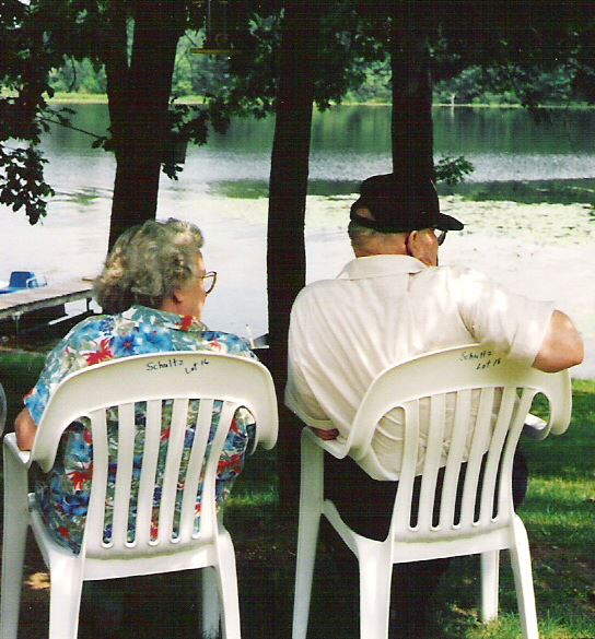 An elderly couple sitting on white plastic chairs by a lake with trees in the background.
