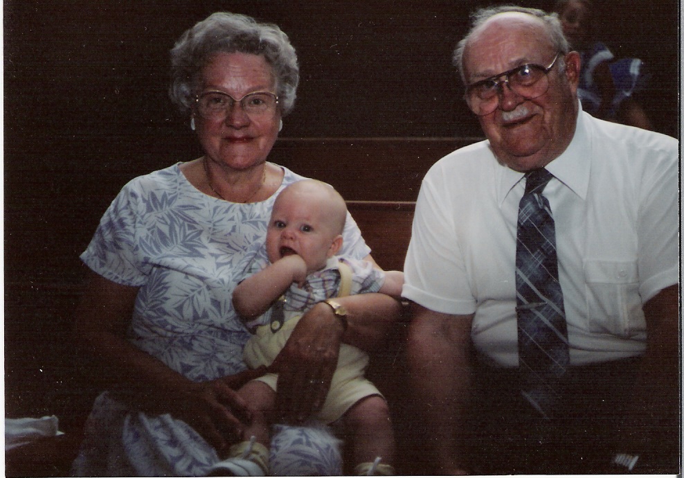 Smiling elderly woman wearing glasses holding a baby with fist in mouth, seated next to a smiling elderly man in glasses and tie.