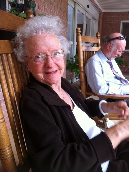 Smiling elderly woman with white curly hair and glasses sitting in a wooden rocking chair on a porch with a man in the background.