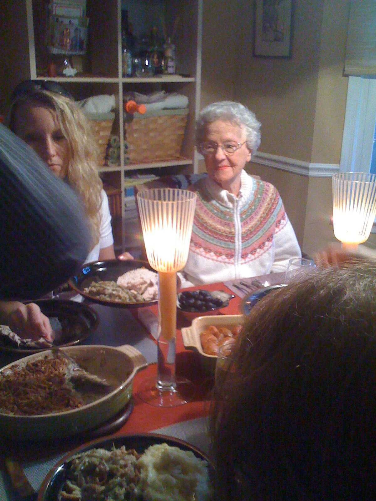Elderly woman and others seated around a dining table with lit candles and plates of food.