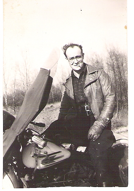 Man wearing a leather jacket and gloves sits on a motorcycle outdoors with leafless trees in the background.