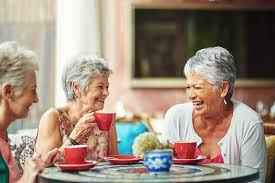Three elderly women with gray hair enjoying conversation and tea together at a round table.