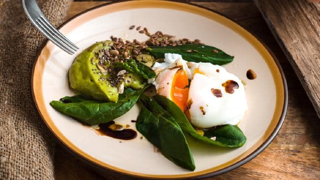 Poached egg with avocado, spinach, sunflower seeds, and flax seeds drizzled with balsamic glaze on a beige plate.