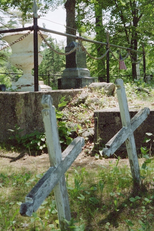 Two aged wooden crosses leaning among grass and plants in a shaded cemetery with headstones and an American flag in the background.