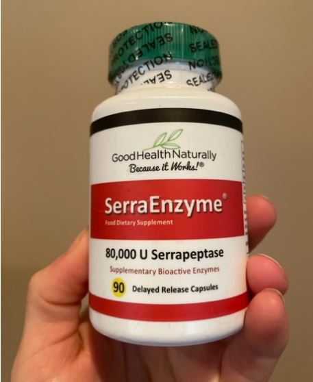 Hand holding a bottle of Good Health Naturally SerraEnzyme dietary supplement with 80,000 U Serrapeptase and 90 delayed release capsules.