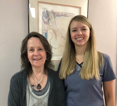 Two smiling women standing indoors in front of a framed abstract line drawing on the wall.