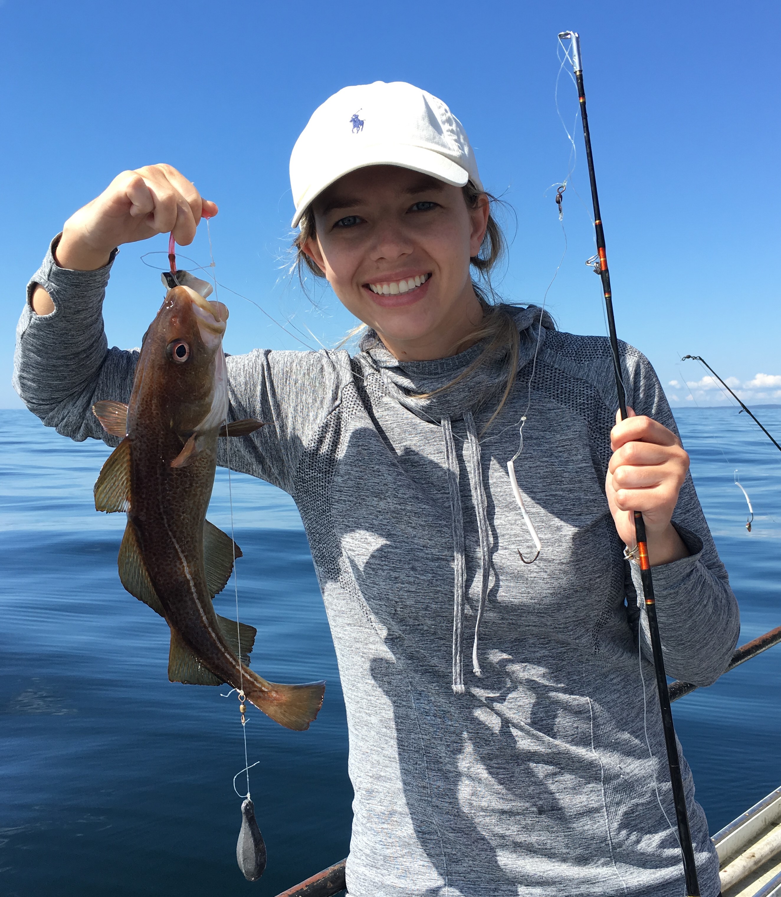 Smiling woman in a white cap and gray hoodie holding a fishing rod and a caught fish on a boat with calm ocean and blue sky in the background.