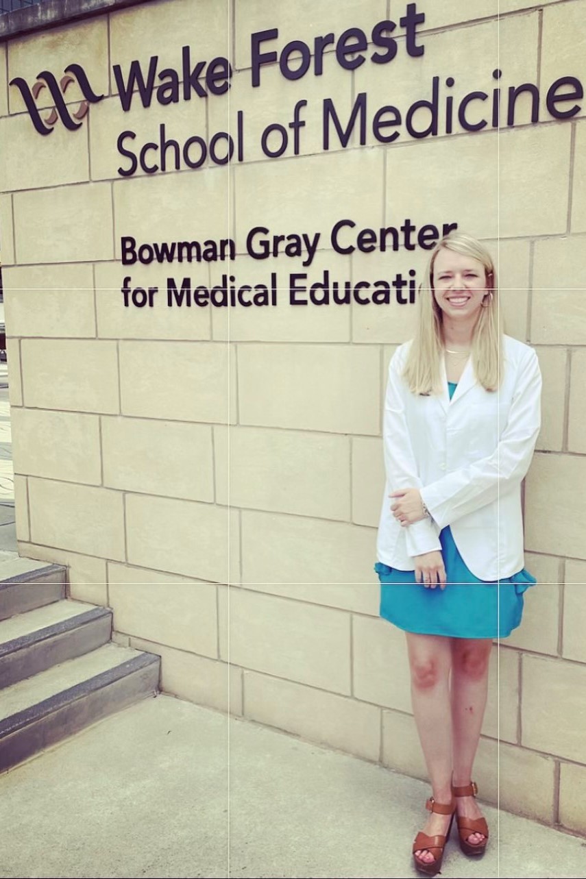 Smiling woman in a white coat and teal dress standing outside the Wake Forest School of Medicine Bowman Gray Center for Medical Education.
