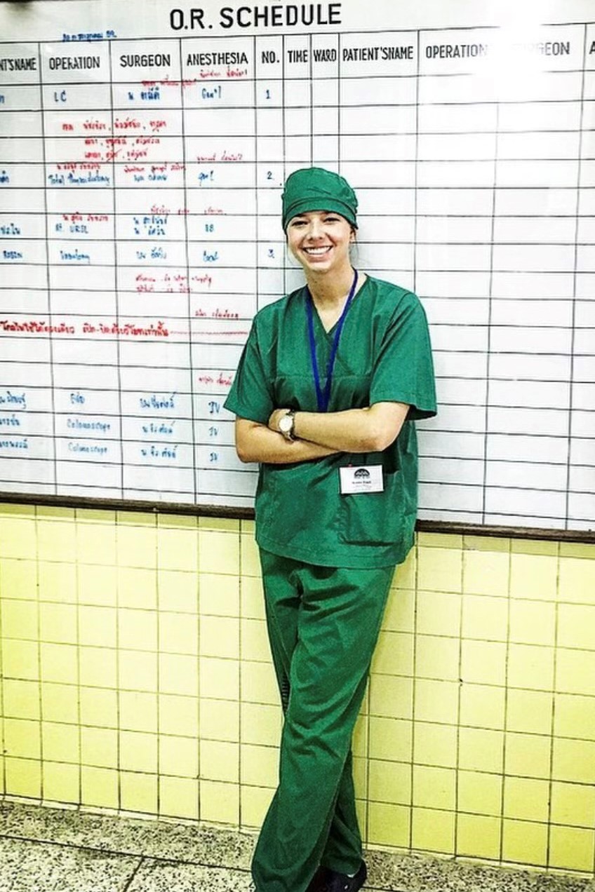 Smiling medical professional in green scrubs and cap standing with arms crossed in front of an operating room schedule board.