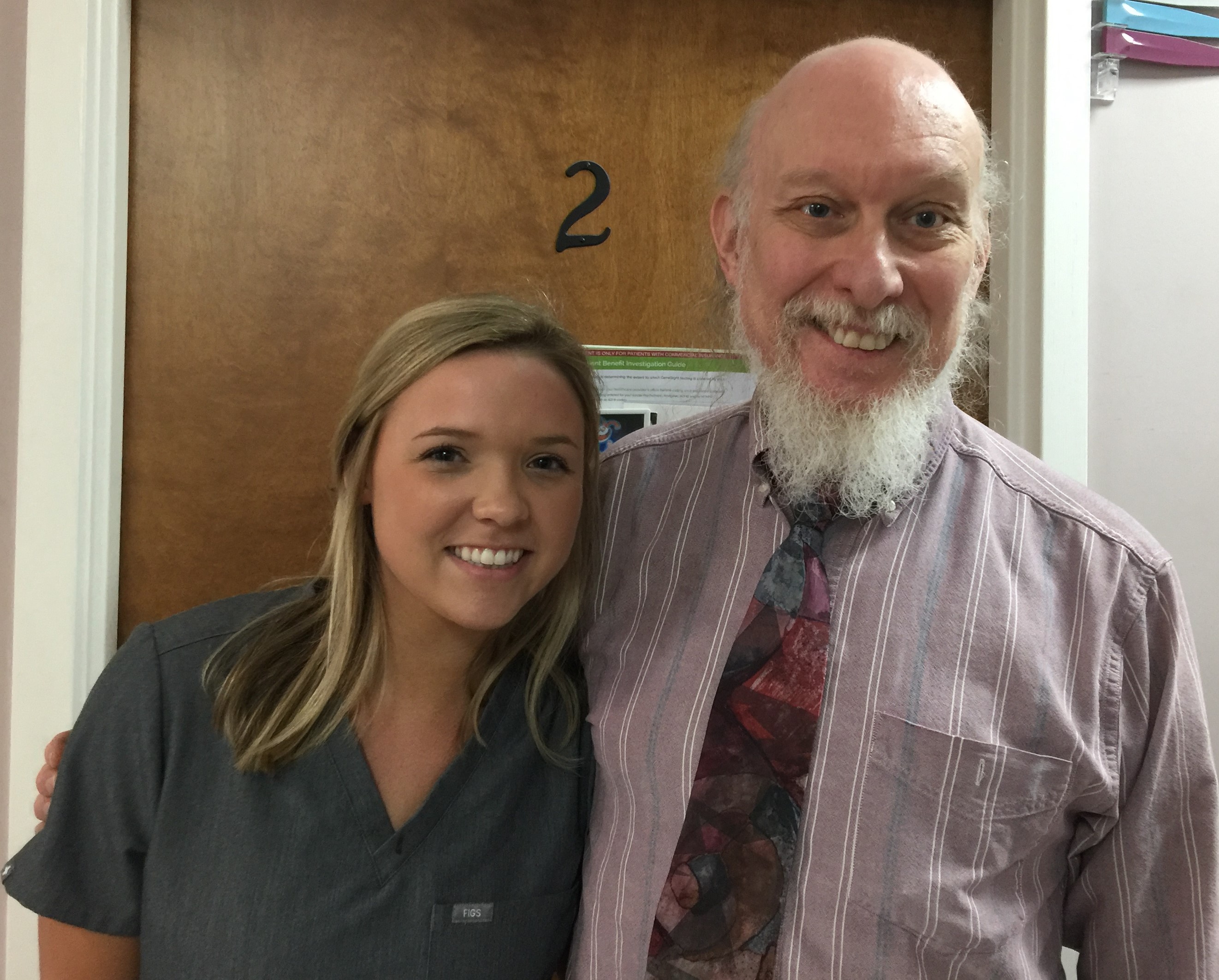 A smiling young woman in gray scrubs stands next to a smiling older man with a white beard wearing a striped shirt and patterned tie in front of a wooden door numbered 2.