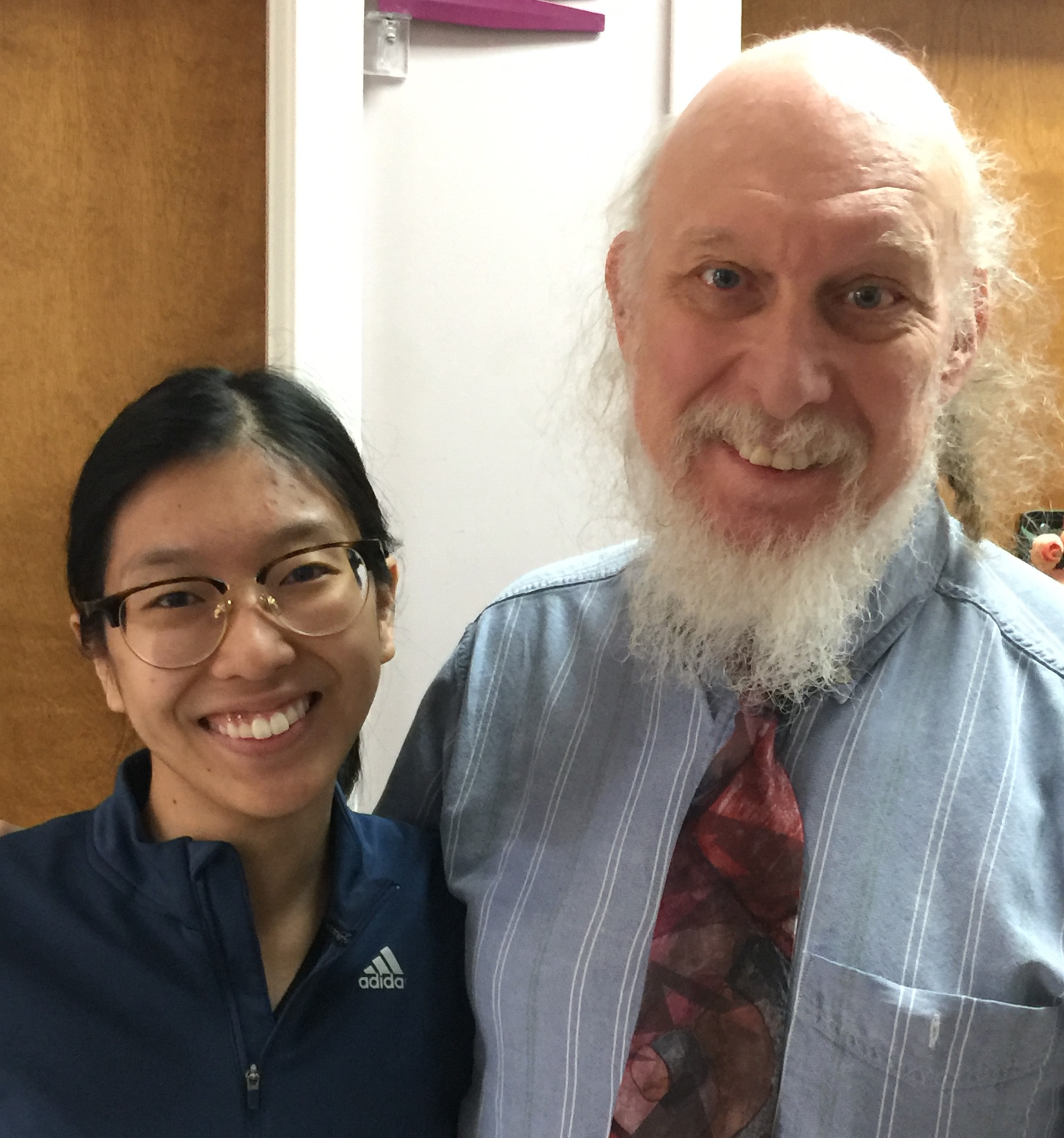 Smiling young woman with glasses and an older man with a white beard wearing a striped shirt and patterned tie, standing indoors.