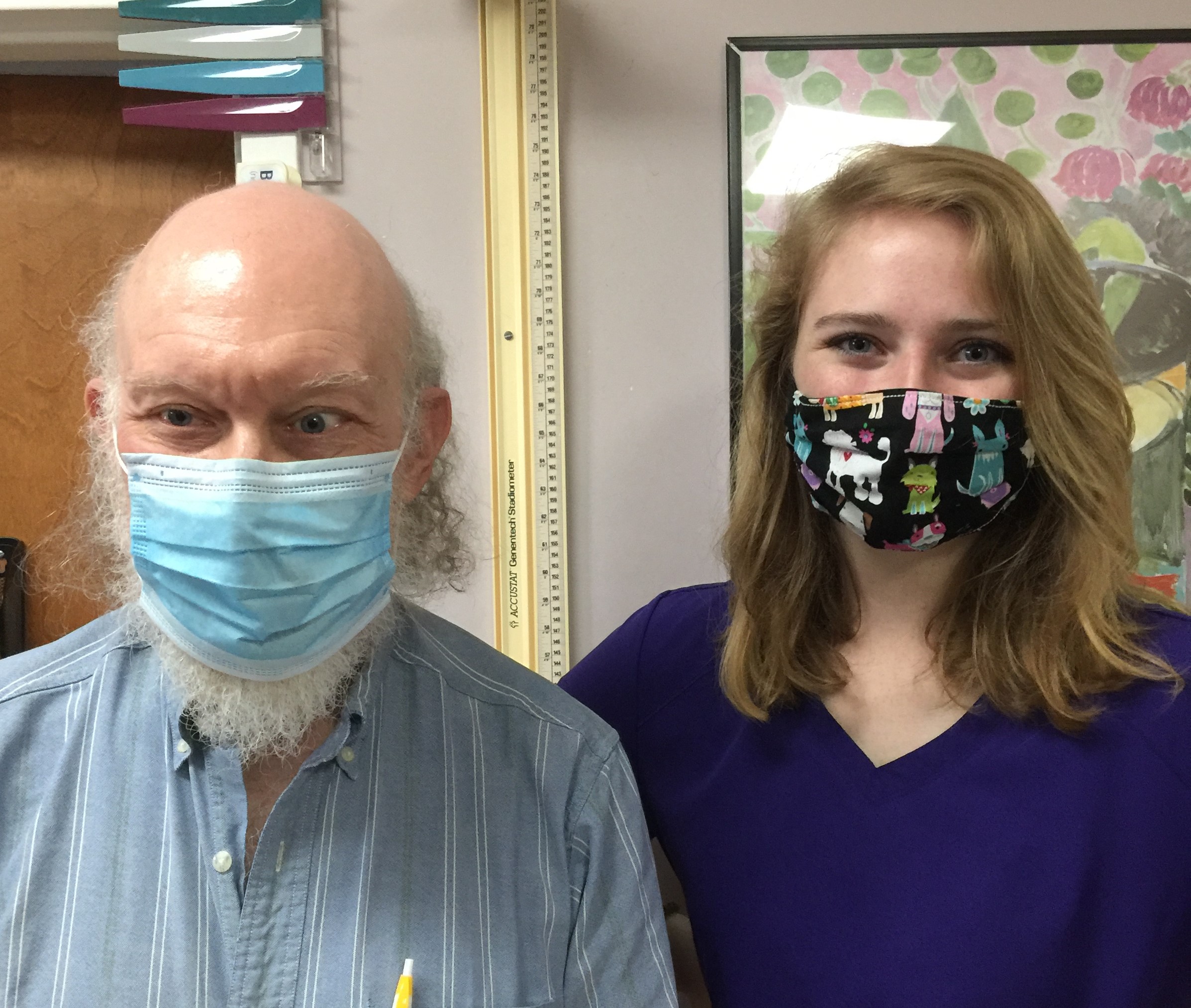 Man with white beard wearing a blue surgical mask and striped shirt stands next to a woman in purple scrubs wearing a colorful dog-patterned face mask.