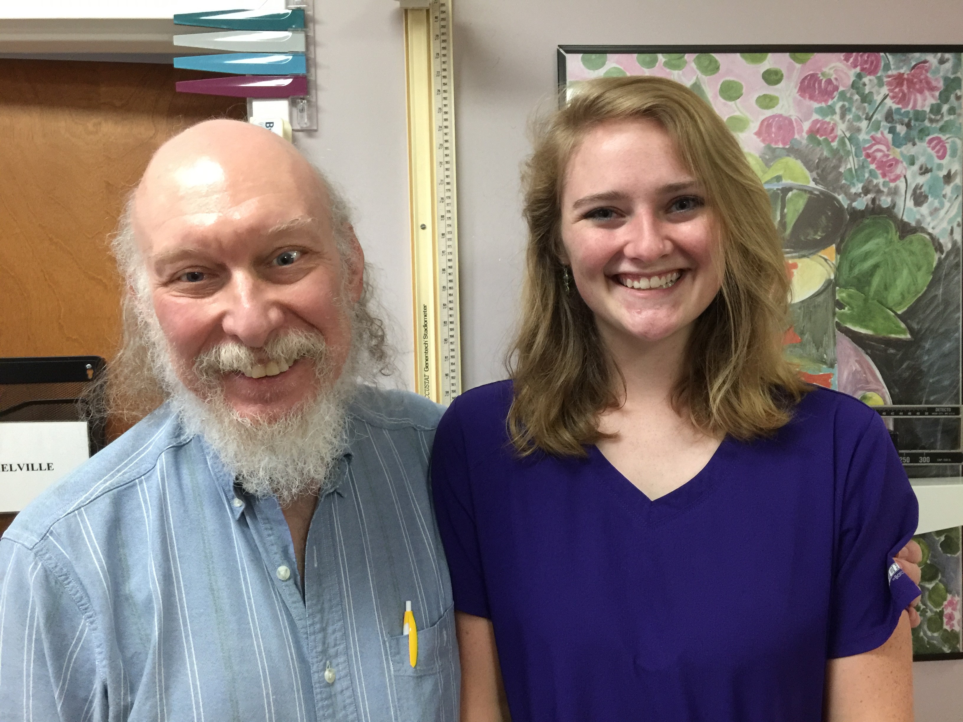 Smiling man with white beard and bald head in blue striped shirt stands next to a smiling young woman with shoulder-length blonde hair wearing a purple top.