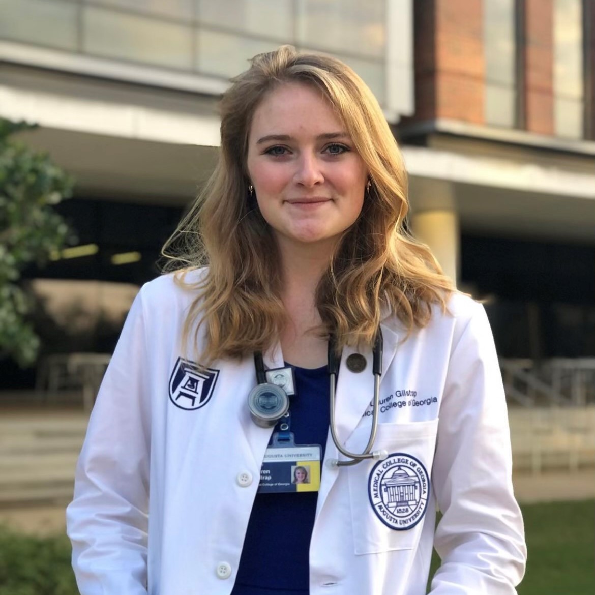 Young woman in a white medical coat with stethoscope, standing outside a building.
