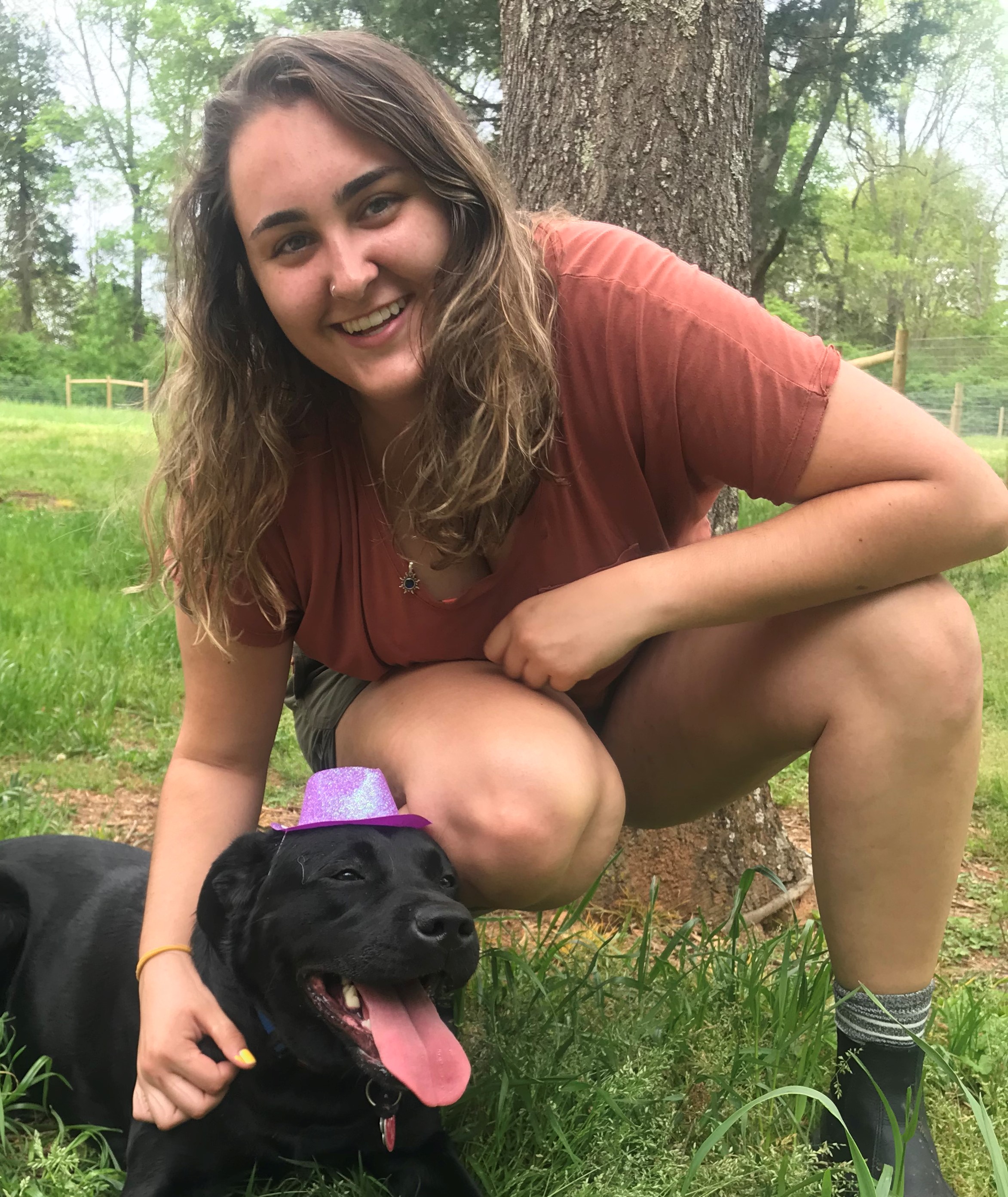 Smiling young woman crouching next to a black dog wearing a small purple glittery hat outdoors.