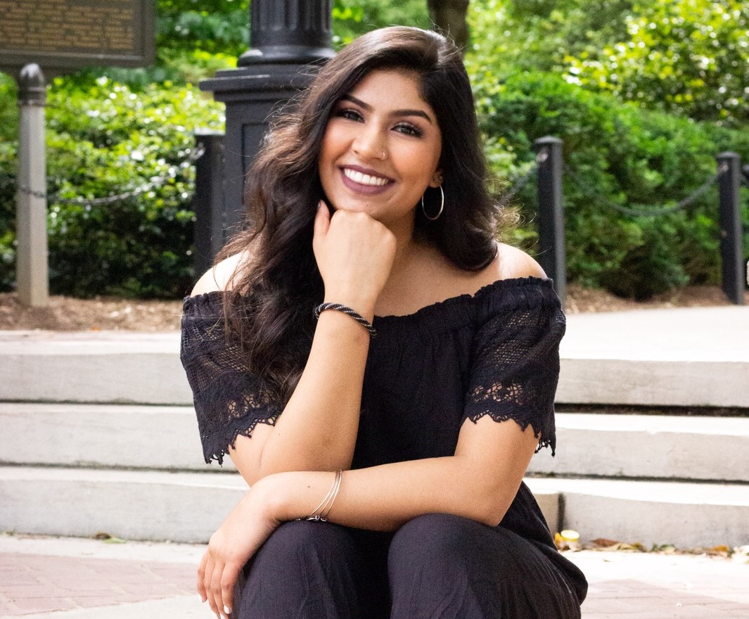 Smiling woman with long dark hair sitting outdoors on steps wearing a black off-shoulder top.