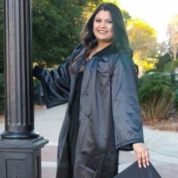 Smiling woman in a black graduation gown holding a cap and leaning against a lamppost outdoors.