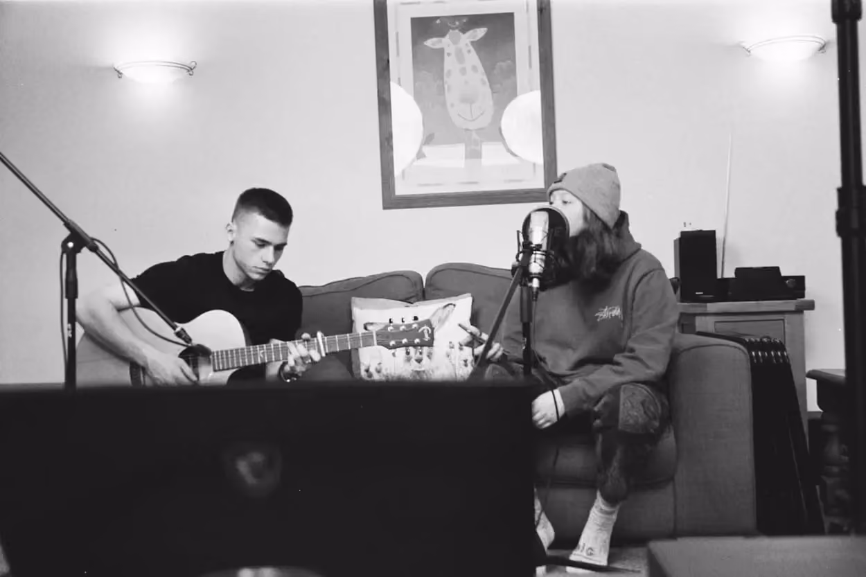 Two men playing acoustic guitars in a cozy living room, surrounded by warm lighting and comfortable furniture. 