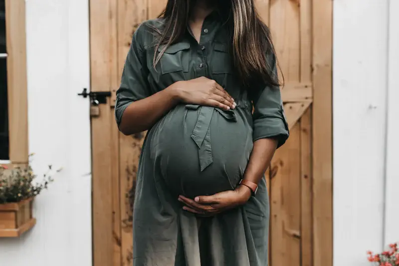 An image of a far along pregnant woman in a dress from the shoulders down, with both hands resting on her stomach.