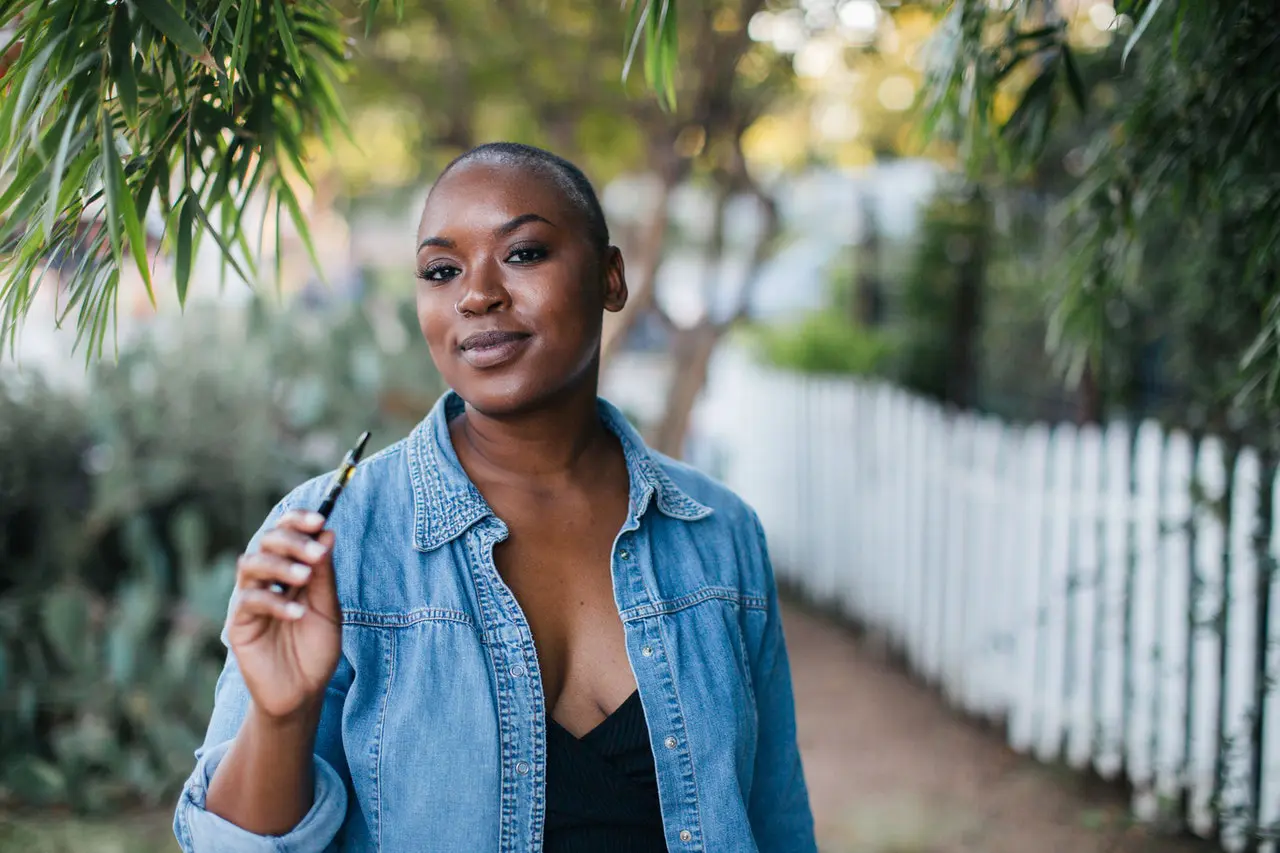 A black woman with a shaved head stands outside holding a vape pen.
