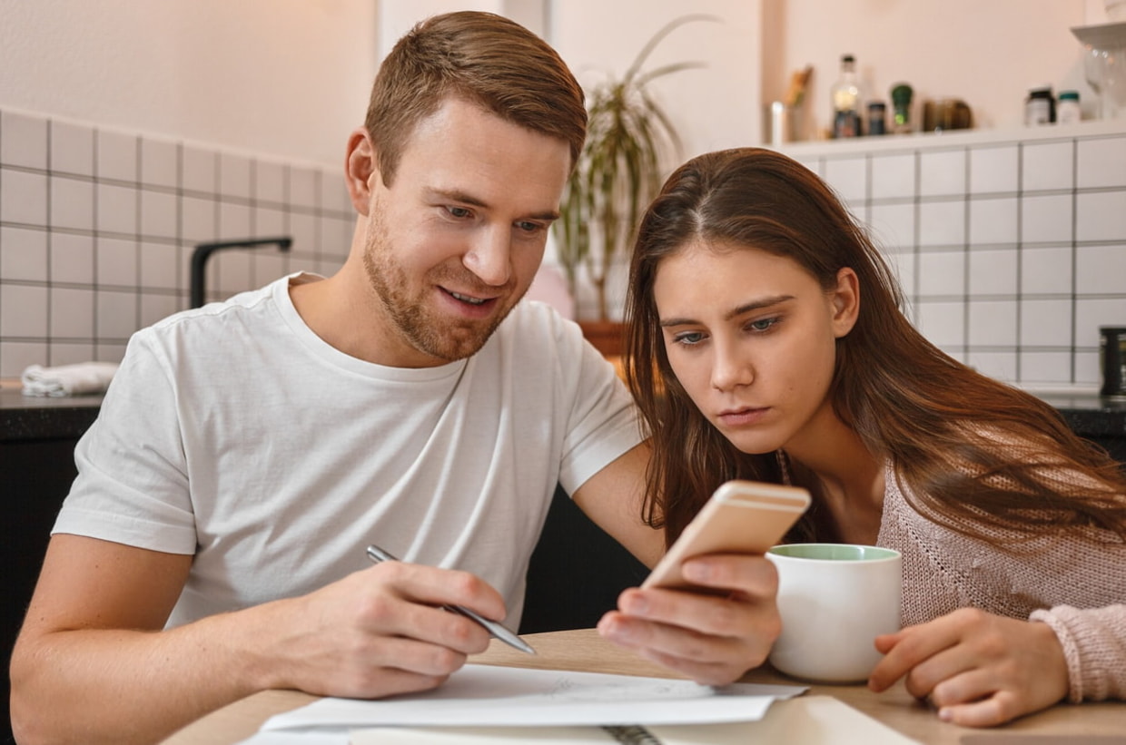 Couple standing in a kitchen, both focused on a smartphone while discussing something together. 