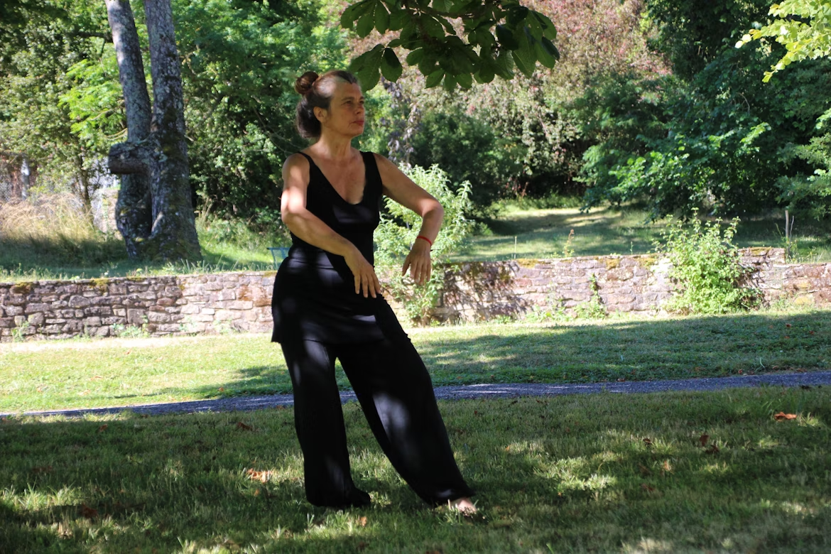 A woman in a black tank top and black pants standing in a grass field doing Tai Chi.