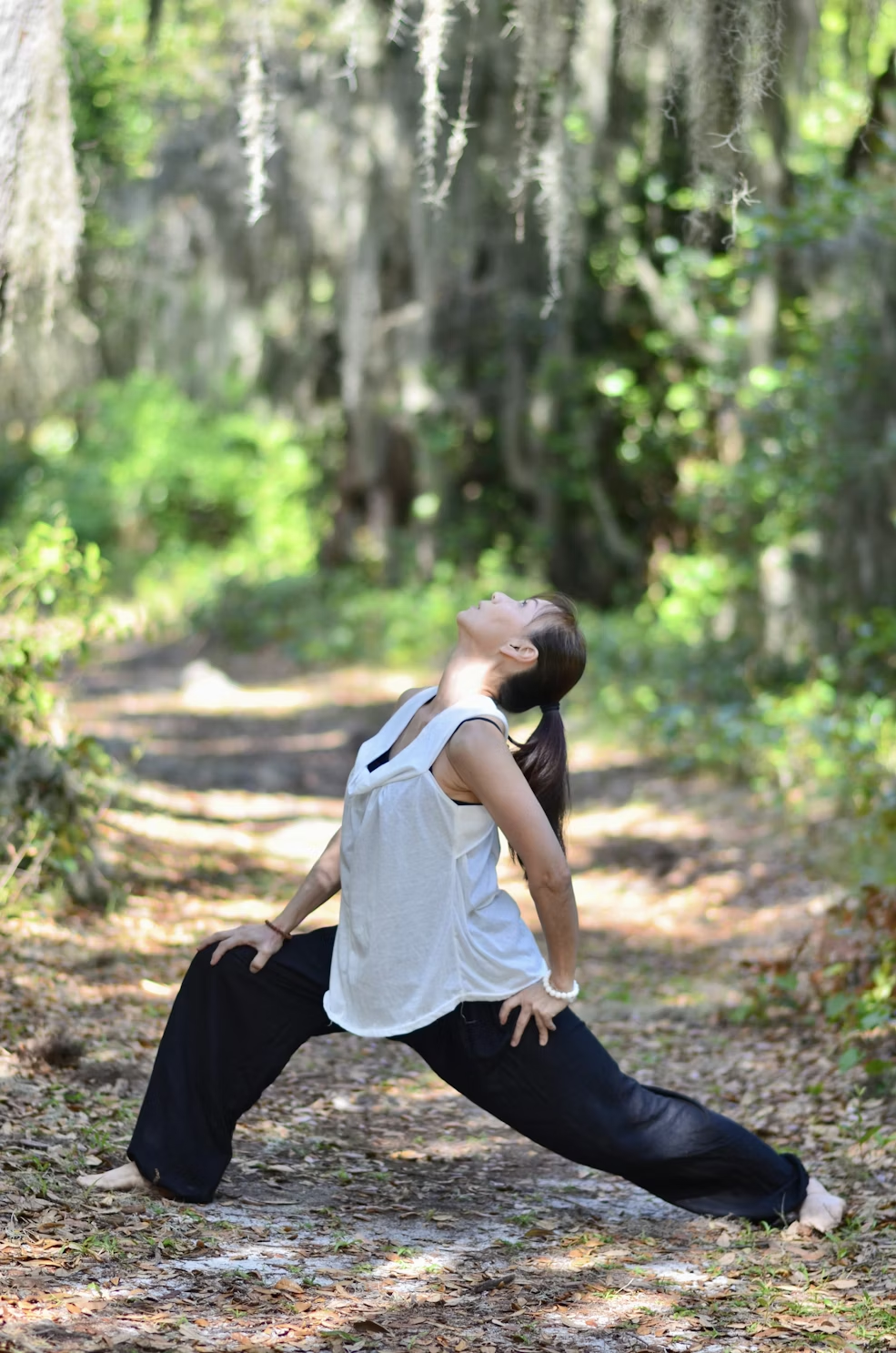 A woman in black pants and a white tank top is stretching in the woods. 