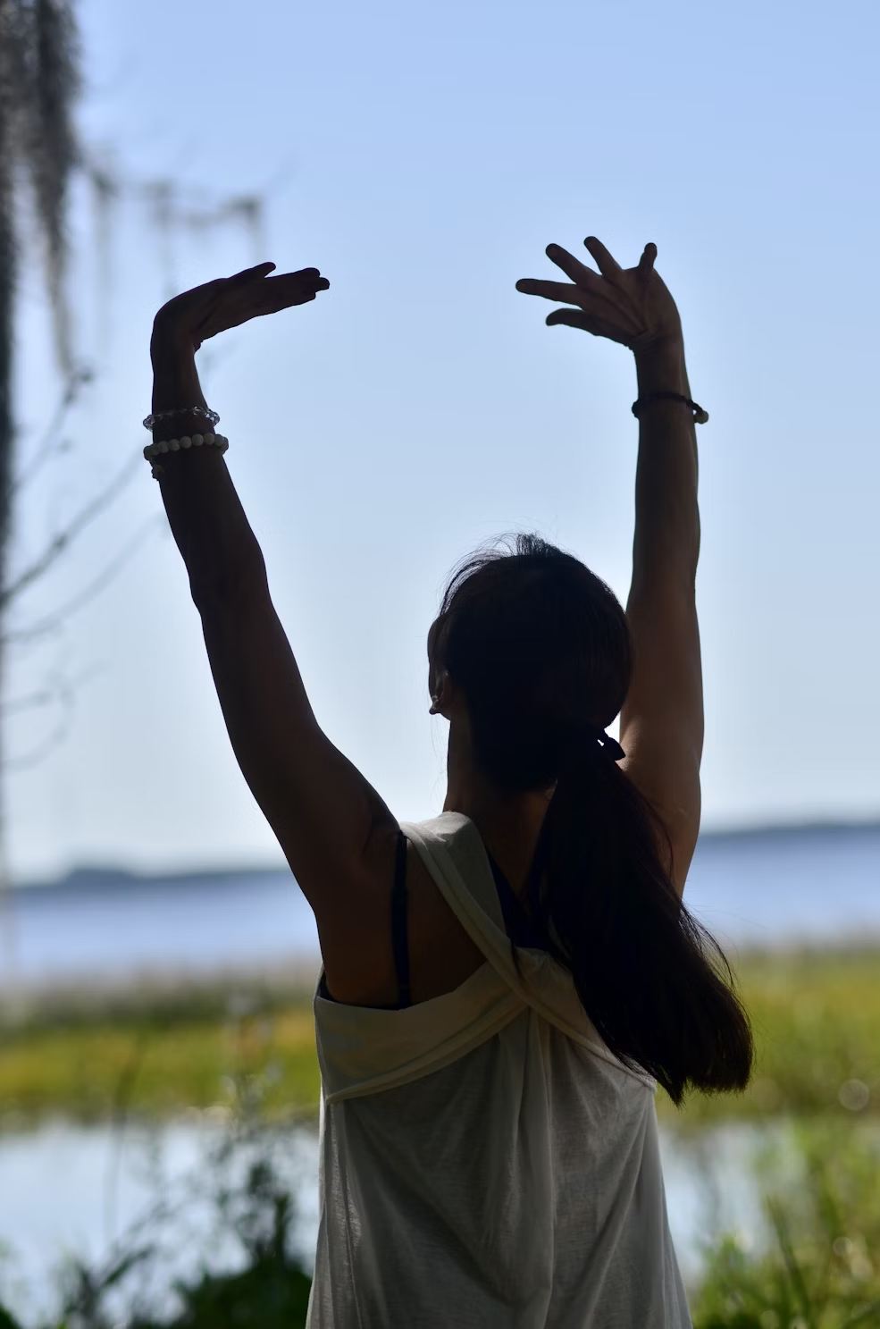 A woman in a white tank top stretching in front of a body of water.