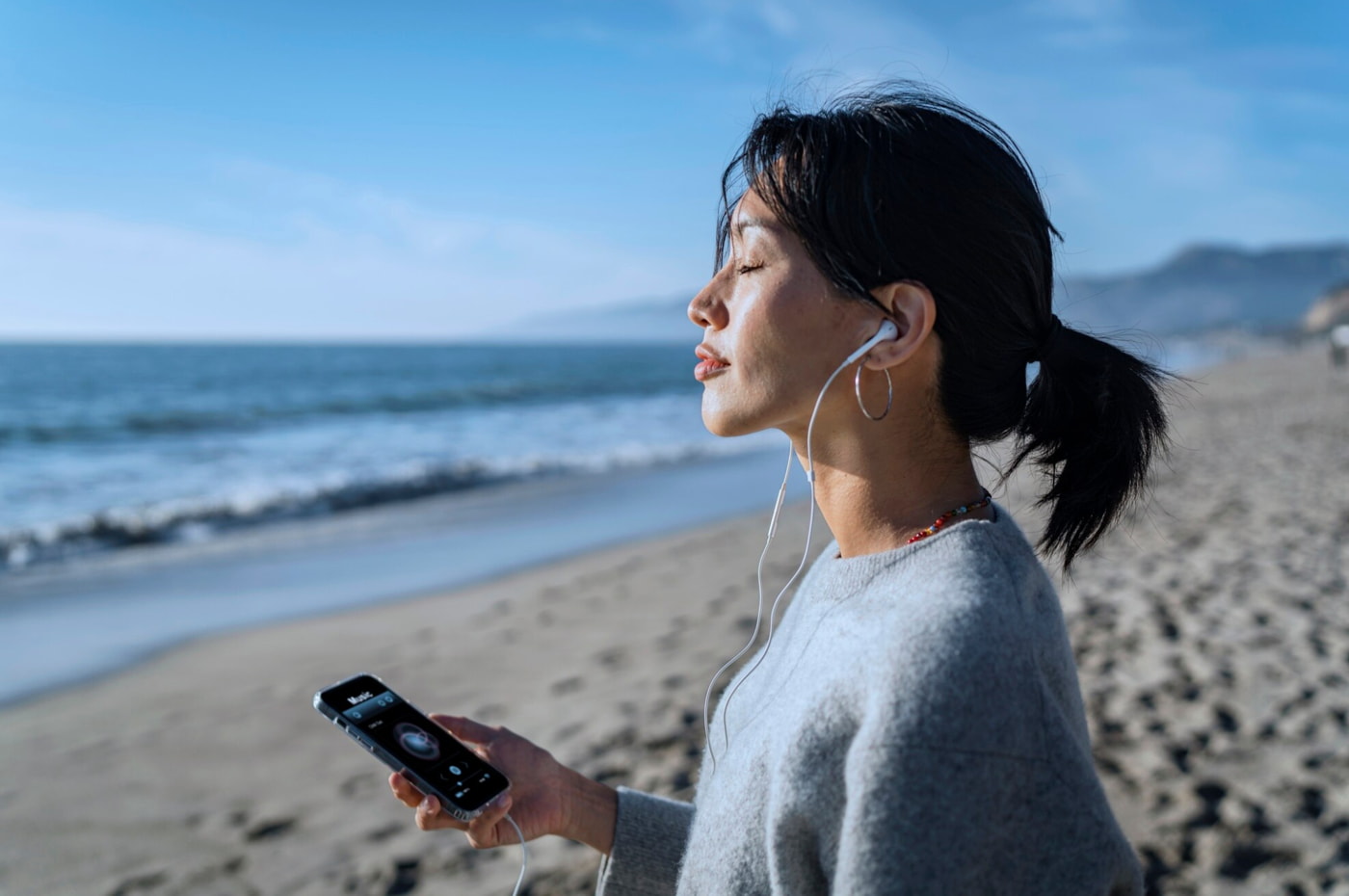 A woman stands on the beach, enjoying music on her phone with the ocean waves in the background. 
