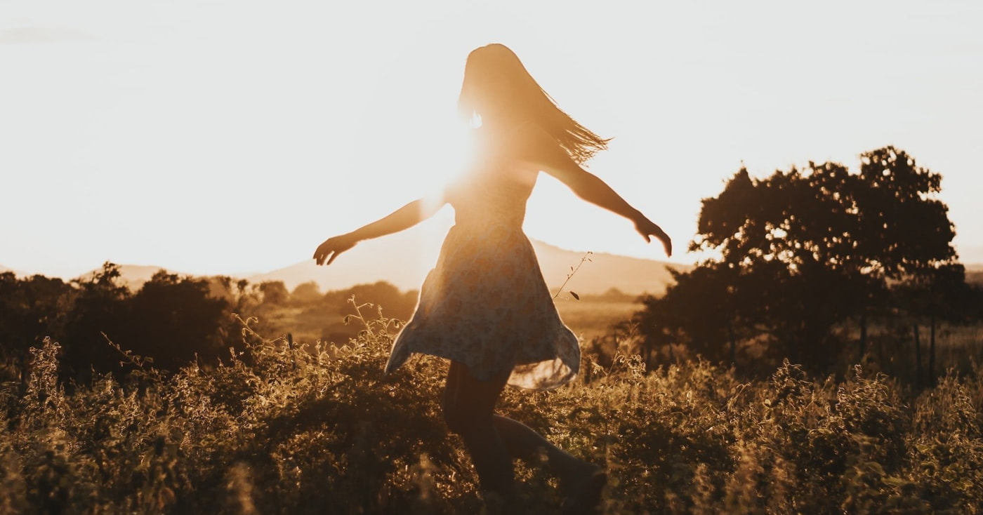 A woman in a flowing dress walks through a field during a vibrant sunset, with warm colors illuminating the scene. 
