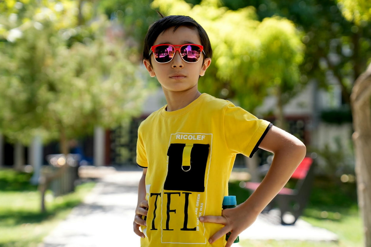 A young boy wearing sunglasses and a bright yellow shirt, smiling and enjoying a sunny day outdoors. 