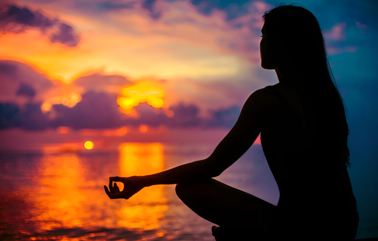 A woman meditates peacefully by the ocean, silhouetted against a vibrant sunset sky. 
