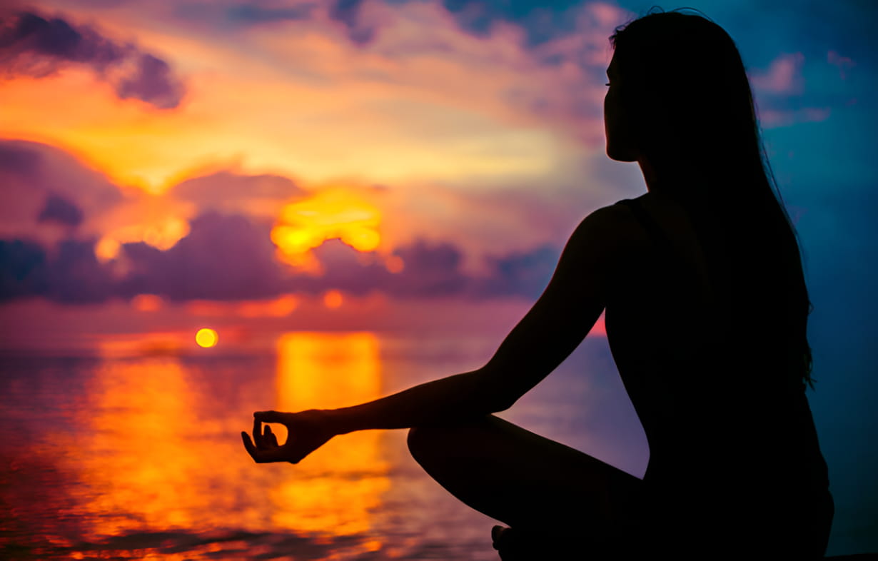 A woman meditates peacefully by the ocean, silhouetted against a vibrant sunset sky. 