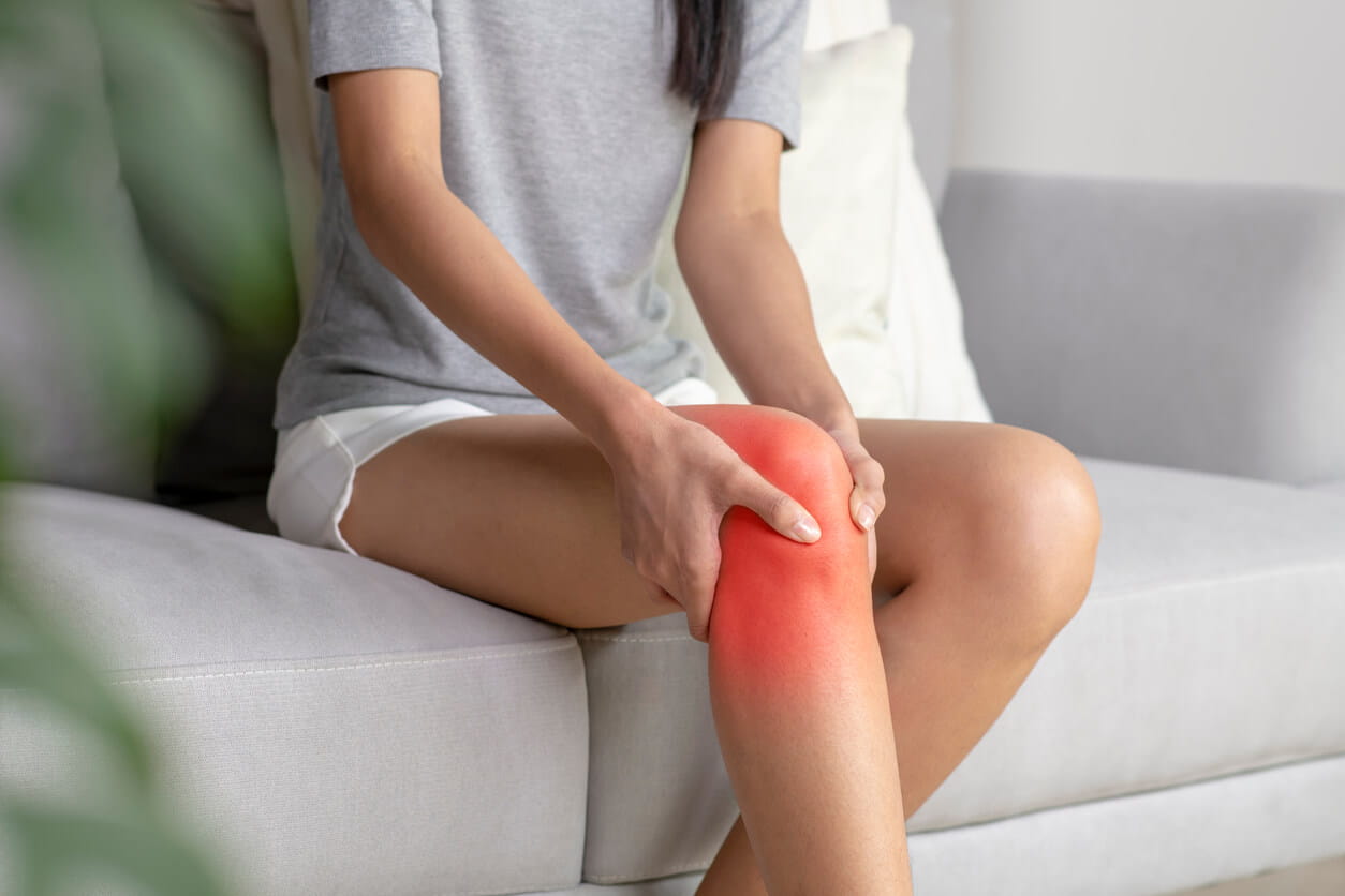 A woman sitting on a couch, holding her knee in pain, with a concerned expression on her face. 