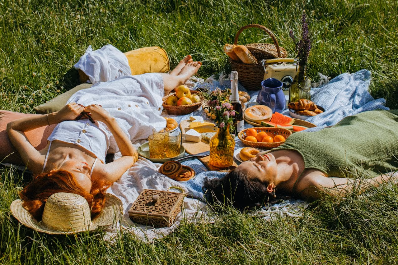 two women lie in a lush, grassy field on a picnic basket laid with fruits and pastries as they enjoy the warmth and prosperity of Litha and the height of summer