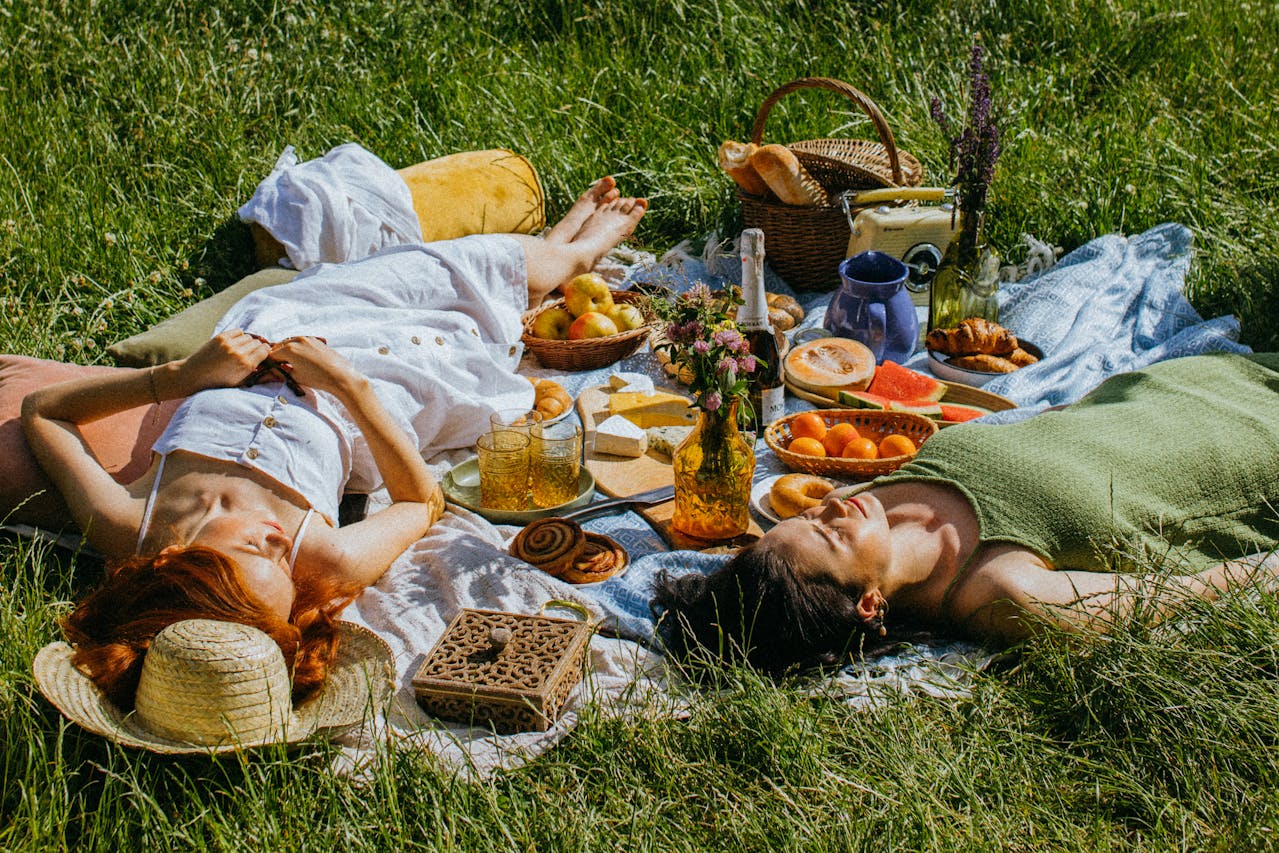two women lie in a lush, grassy field on a picnic basket laid with fruits and pastries as they enjoy the warmth and prosperity of Litha and the height of summer