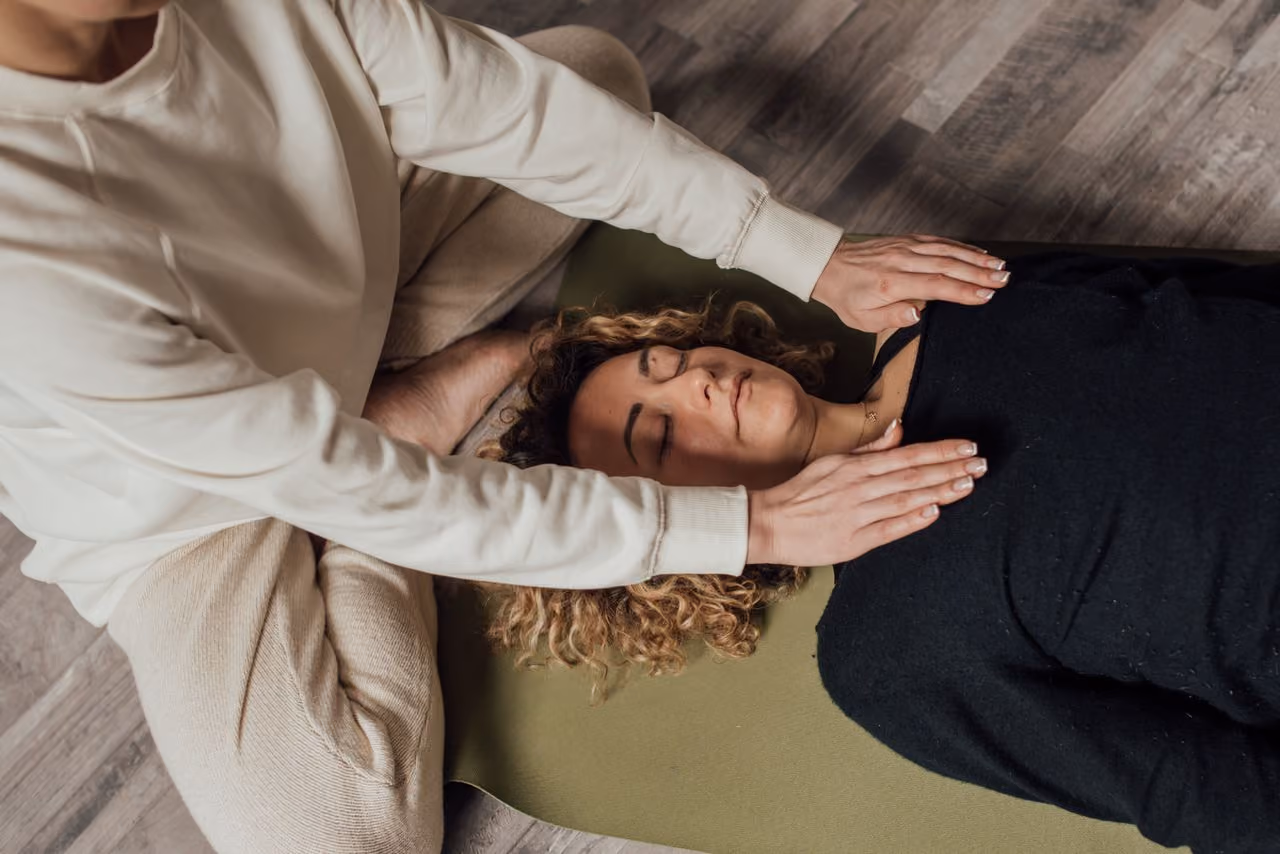 woman wearing sweatpants lying on her back on a yoga mat while a reiki practitioner places her hands lightly on the woman's shoulders, sensing energy flow in the body